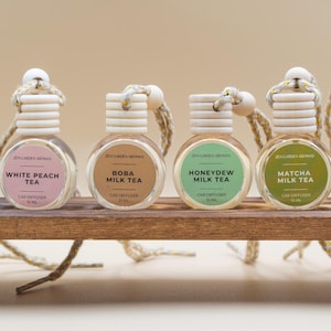 May include: Six car air fresheners with different flavors, including Thai Tea, White Peach Tea, Boba Milk Tea, Honeydew Milk Tea, Matcha Milk Tea, and Taro Milk Tea. Each bottle is clear with a wooden cap and a label. They are displayed on a small wooden shelf.