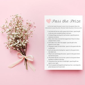 May include: A pink card with a heart at the top and the text "Pass the Prize" in black. The card has instructions for a game where guests pass a prize around the room.
