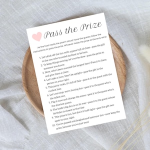 May include: A white card with a pink heart and the text "Pass the Prize" in black. The card contains instructions for a game where guests pass a prize around the room based on different criteria.