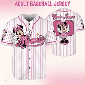 May include: White baseball jersey with pink pinstripes, featuring a Minnie Mouse graphic. The front displays "Minnie" in pink script, while the back has a pink number and space for a name. The sleeves have Minnie Mouse graphics.