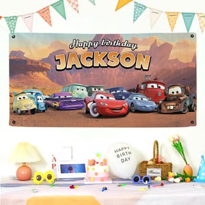 May include: A birthday banner featuring the text "Happy birthday JACKSON" and cartoon car characters. The scene includes a table with a cake, balloons, and party decorations, with bunting overhead.