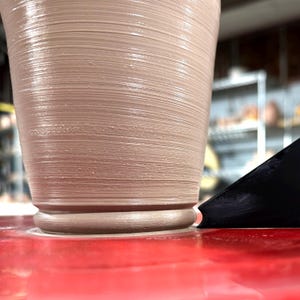 K&ouml;nnte beinhalten: Nahaufnahme einer T&ouml;pferscheibe mit einer Tonvase, die geformt wird. Die Vase ist hellbraun und hat eine glatte, gleichm&auml;&szlig;ige Oberfl&auml;che. Ein schwarzes Werkzeug wird verwendet, um den Ton zu formen.