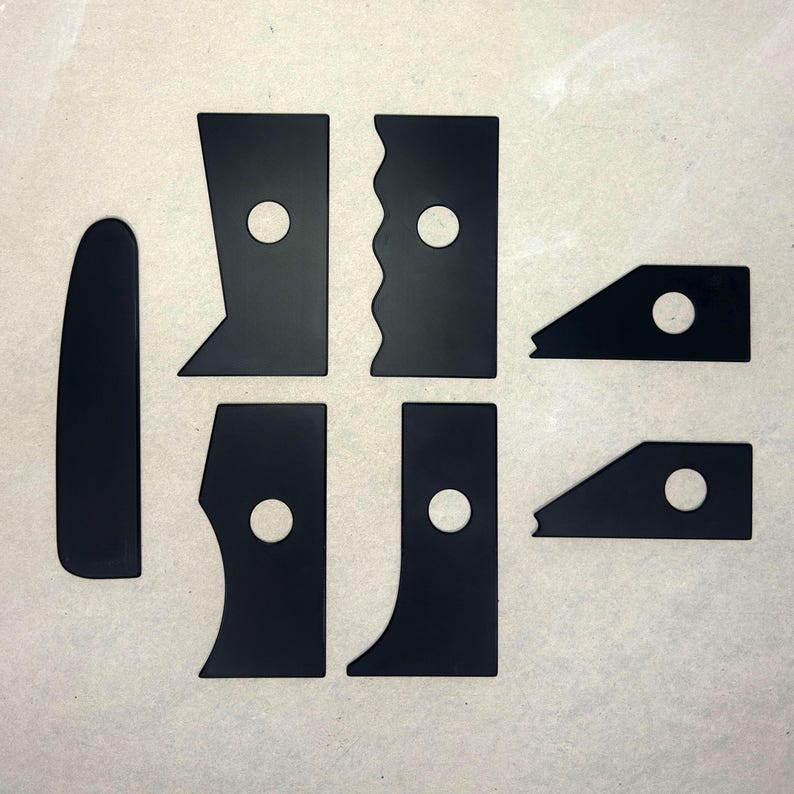 May include: Seven black plastic tools with rounded edges and holes in the center. The tools are arranged in a grid pattern on a light gray surface.