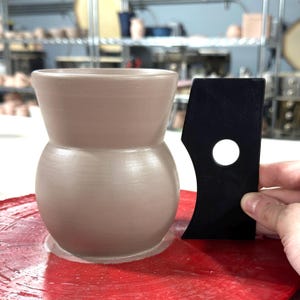 K&ouml;nnte beinhalten: Eine hellbraune Keramikvase mit abgerundetem Boden und einer breiten &Ouml;ffnung. Die Vase wird auf einer T&ouml;pferscheibe mit einem schwarzen Plastikwerkzeug geformt.