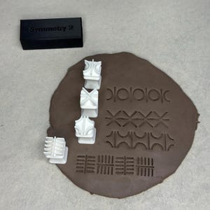 Puede incluir: Una colección de sellos de arcilla blanca y un contenedor rectangular negro con la etiqueta "Symmetry 2" se muestran sobre una superficie de arcilla marrón. Los sellos tienen varios diseños geométricos, y la arcilla muestra impresiones de los patrones.