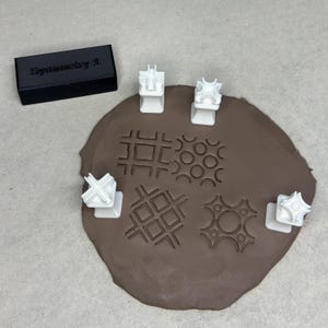 May include: A black rectangular object with the text "Symmetry 1." is next to a brown clay shape with various geometric patterns pressed into it. White plastic tools are positioned around the clay, showcasing the patterns they create.