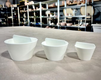 May include: Three white ceramic bowls with handles, arranged in order of size from largest to smallest.