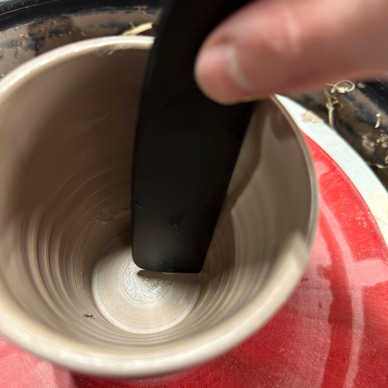 K&ouml;nnte beinhalten: Nahaufnahme einer T&ouml;pferscheibe mit einer Keramiktasse, die geformt wird. Ein schwarzes Werkzeug wird verwendet, um den Ton zu gl&auml;tten.