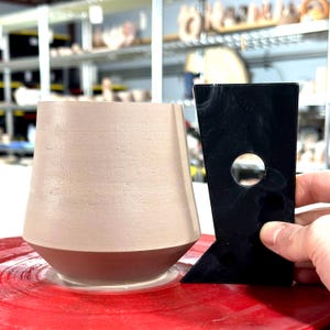 K&ouml;nnte beinhalten: Eine hellbraune Keramiktasse mit abgerundetem Boden und breitem Rand wird auf einer T&ouml;pferscheibe geformt. Ein schwarzes Plastikwerkzeug mit einem Loch in der Mitte wird verwendet, um die Tasse zu formen.