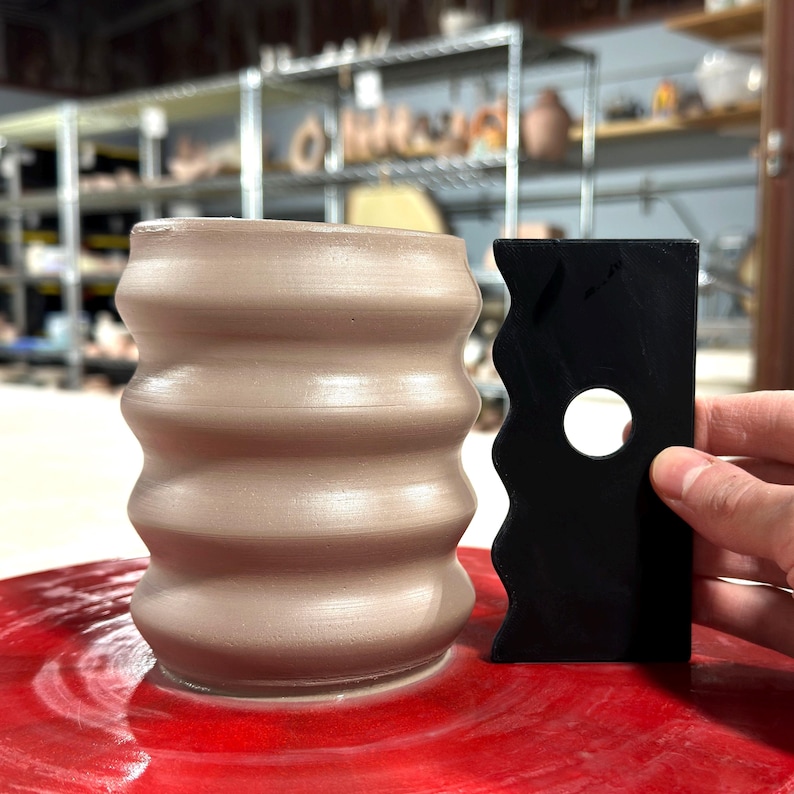 K&ouml;nnte beinhalten: Eine hellbraune Keramiktasse mit einem welligen, strukturierten Design wird auf einer T&ouml;pferscheibe geformt. Ein schwarzes Plastikwerkzeug mit einer wellenf&ouml;rmigen Kante und einem runden Loch wird verwendet, um die Tasse zu formen.
