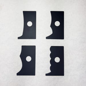 Puede incluir: Cuatro herramientas de costilla de cerámica negras con diferentes formas de borde. Cada herramienta tiene un pequeño agujero central. Las herramientas están dispuestas sobre un fondo blanco, mostrando sus variados perfiles para moldear arcilla.