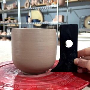K&ouml;nnte beinhalten: Eine hellbraune Keramiktasse auf einer roten T&ouml;pferscheibe mit einem schwarzen Messwerkzeug daneben.