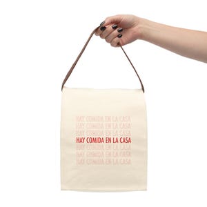 May include: A white canvas tote bag with brown straps. The bag has the text "HAY COMIDA EN LA CASA" repeated multiple times in red and pink.