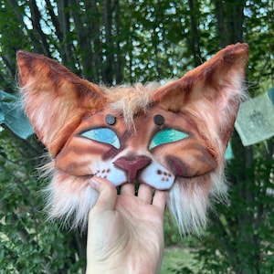 Può includere: Una maschera felina artigianale in pelliccia sintetica arancione e marrone, con grandi orecchie a punta e occhi di paillettes color turchese brillante. La maschera presenta un naso scolpito, muso bianco con macchie marroni e finitura morbida.