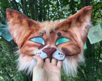 May include: A handcrafted feline mask featuring orange and brown faux fur, large pointed ears, and bright teal sequined eyes. The mask includes a sculpted nose, white muzzle detailing with brown spots, and a soft, textured finish.