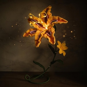 Pode incluir: Uma lâmpada de flor de lírio em crochê com grandes pétalas amarelas com detalhes roxos e um botão amarelo menor num caule verde. O centro da flor grande é iluminado por fios finos e flexíveis de luz LED que emitem um brilho quente.