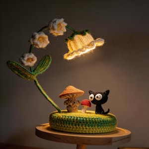 May include: A handcrafted crochet lamp featuring a lily of the valley design with a glowing yellow lampshade. The base includes a mushroom and a black cat figurine. The lamp is set on a wooden tray.