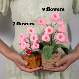 May include: Two crochet flower arrangements. One pot has 7 pink flowers, the other has 9. The flowers are in brown crochet pots with green leaves. One pot has a red mushroom.