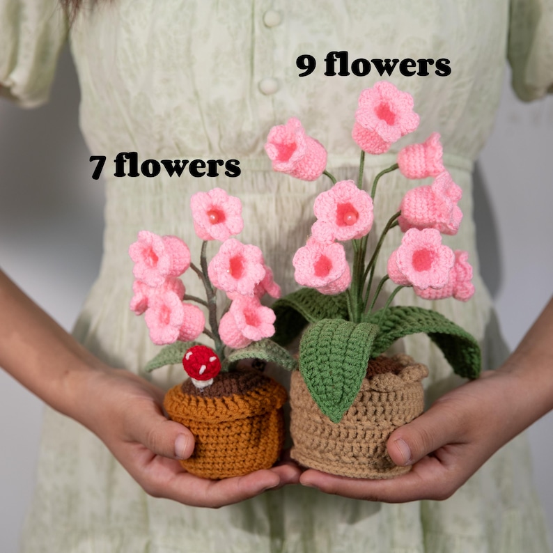 May include: Two crochet flower arrangements. One pot has 7 pink flowers, the other has 9. The flowers are in brown crochet pots with green leaves. One pot has a red mushroom.
