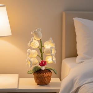 May include: A handcrafted crochet lamp shaped like lily of the valley flowers. The white flowers are illuminated with warm light, and the lamp includes a small red mushroom. The lamp is in a brown crochet pot, sitting on a white nightstand.