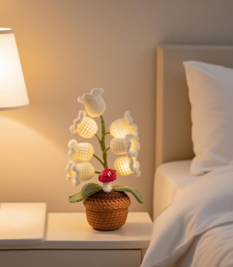 May include: A handcrafted crochet lamp shaped like lily of the valley flowers. The white flowers are illuminated with warm light, and the lamp includes a small red mushroom. The lamp is in a brown crochet pot, sitting on a white nightstand.