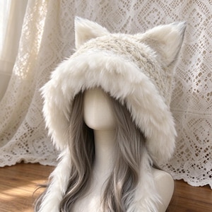 May include: A cream-colored faux fur hood featuring two prominent, pointed cat-like ears on top. The hood is lined with thick, soft white fur trim along the face opening and includes long, dangling fur-trimmed side panels that drape downward.