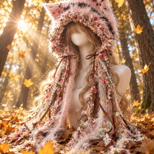 May include: A fuzzy pink cat-ear hood featuring a textured, multi-material trim with dangling ribbons, lace, and small floral accents. The hood is crafted from soft, shaggy yarn and includes long, decorative fringe elements that drape down the sides.