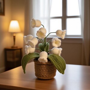 May include: A handcrafted crochet lily of the valley plant in a woven basket. The white bell-shaped flowers are clustered on green stems with green leaves. The basket is a light brown color, and the plant sits on a wooden table.
