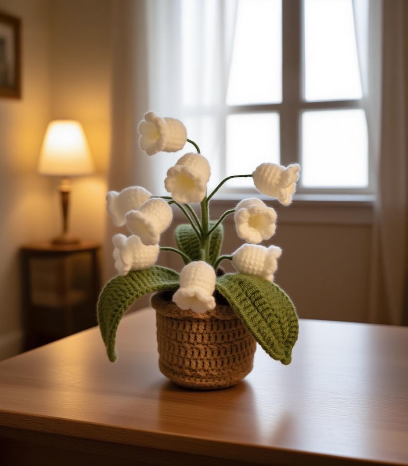 May include: A handcrafted crochet lily of the valley plant in a woven basket. The white bell-shaped flowers are clustered on green stems with green leaves. The basket is a light brown color, and the plant sits on a wooden table.