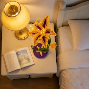 May include: A crocheted flower lamp featuring yellow and purple petals with integrated warm LED fairy lights. The flower is attached to a green stem and a circular purple base decorated with a yellow star. An open book lies next to the lamp.