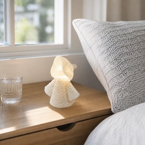 May include: A small, cream-colored, crocheted figure with a hood and wide sleeves, illuminated from within to emit a soft, warm glow. The textured, yarn-based object stands upright, featuring a simple, rounded silhouette and a hollow interior.