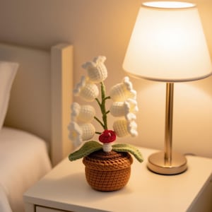 May include: A handcrafted crochet lily of the valley plant in a brown pot. The white flowers are clustered on a green stem, with green leaves and a red mushroom accent. The plant sits on a white nightstand next to a lamp.
