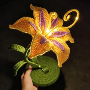 May include: A handmade crocheted flower lamp. The flower has yellow and purple petals, green leaves, and a green base. The lamp features a glowing centre and a curly stem with small lights. The design is whimsical and decorative.