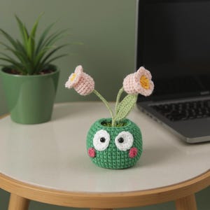 May include: A handmade crochet flower arrangement featuring two pink and yellow flowers with green stems and leaves. The flowers are in a green pot with white eyes and pink cheeks. A small potted plant and a laptop are in the background.