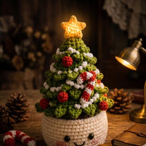以下が含まれることがあります： 光る黄色い星を頂点に飾った緑色の編みぐるみのクリスマスツリー。赤いオーナメント、白いガーランド、赤と白のキャンディケインが装飾されています。ツリーは、黒い目とピンクの頬をした笑顔の顔が描かれたクリーム色の編みぐるみの鉢に入っています。