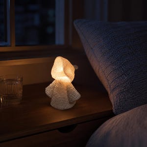 May include: A small, cream-colored, crocheted figure shaped like a hooded character emitting a warm, soft internal glow. The textured, yarn-based structure features a distinct hood and flared sleeves, standing upright as a decorative light source.