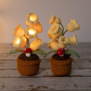May include: Two crochet flower lamps in brown pots. The yellow flowers are lit, with green leaves and a red mushroom accent. The lamps are on a light-colored wooden surface.
