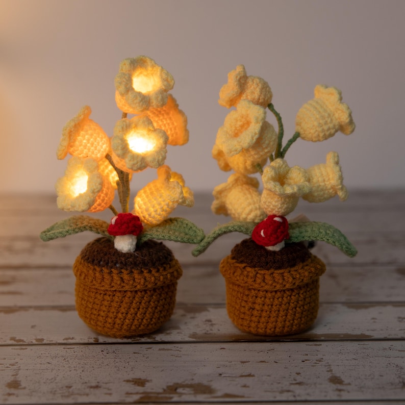 May include: Two crochet flower lamps in brown pots. The yellow flowers are lit, with green leaves and a red mushroom accent. The lamps are on a light-colored wooden surface.
