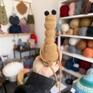 以下が含まれることがあります： 黒と白の目を持つ、手作りのベージュ色の編みぐるみのイモムシのおもちゃ。 イモムシは積み重ねられた丸いセグメントでできており、長い糸の脚が付いています。 おもちゃは手で持たれており、木製のベースが付いています。