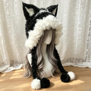 Può includere: Un cappello di peluche con orecchie da animale in pelliccia sintetica nera e bianca. Il cappello presenta grandi orecchie a punta, un bordo spesso in pelliccia bianca attorno al viso e due lunghi lembi laterali con pompon neri e bianchi.