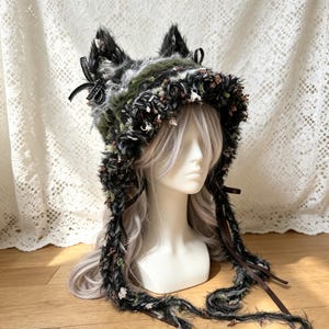 以下が含まれることがあります： 黒、グレー、オリーブグリーンの質感のある毛糸で作られた、猫耳付きのダークトーンのボンネット。帽子には小さな黒いリボンがあしらわれ、両サイドには長く垂れ下がった編み込みの紐が付いています。