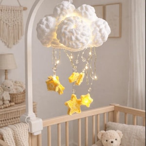 May include: A white cloud-shaped baby mobile with yellow star accents hangs above a wooden crib. The mobile features a soft, textured design with integrated lights and dangling stars. The crib contains a teddy bear and a pillow.