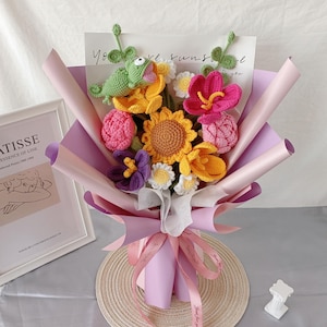 May include: A colorful bouquet of crocheted flowers, including sunflowers, daisies, and other blooms, wrapped in pink and purple paper. A green crocheted frog sits atop the arrangement. The bouquet is tied with pink ribbons.