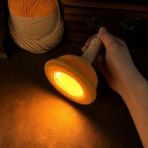 May include: A handheld light fixture featuring a circular, bright orange crocheted shade with a textured surface. The light emits a warm, glowing yellow illumination from the centre, while the handle is wrapped in a neutral-toned, woven cord.