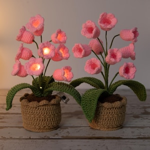 May include: Two crochet flower arrangements with pink illuminated blossoms. The flowers are in brown pots with green leaves. The pots are made of a tan crochet material. The flowers have a warm glow.