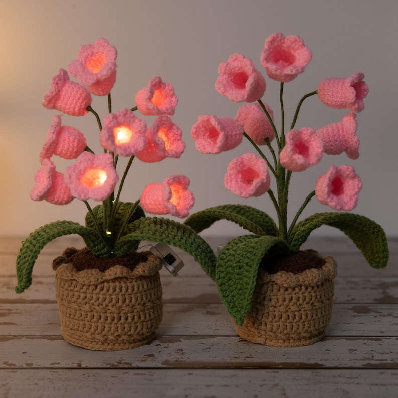 May include: Two crochet flower arrangements with pink illuminated blossoms. The flowers are in brown pots with green leaves. The pots are made of a tan crochet material. The flowers have a warm glow.