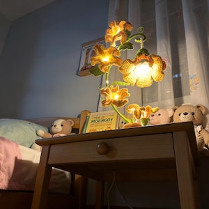 May include: A handcrafted crochet flower lamp with yellow and green details. The lamp features multiple flower-shaped shades with warm lighting, sitting on a wooden nightstand. A book with the text "MOENGGOY" is visible.