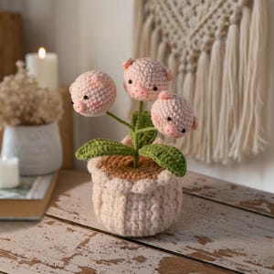 Puede incluir: Un arreglo floral de ganchillo caprichoso con tres flores en forma de cerdos en un tono rosa claro, con pequeñas orejas y hocicos. Las flores están en una maceta rosa claro con un borde festoneado y hojas verdes. El arreglo está sobre una superficie de madera.