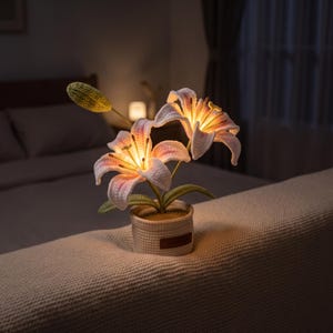 May include: A crocheted lily flower lamp featuring two open white and pink blossoms and one green bud. The flowers are illuminated from within, emitting a warm glow. The plant is set in a small, textured, cylindrical beige woven pot.