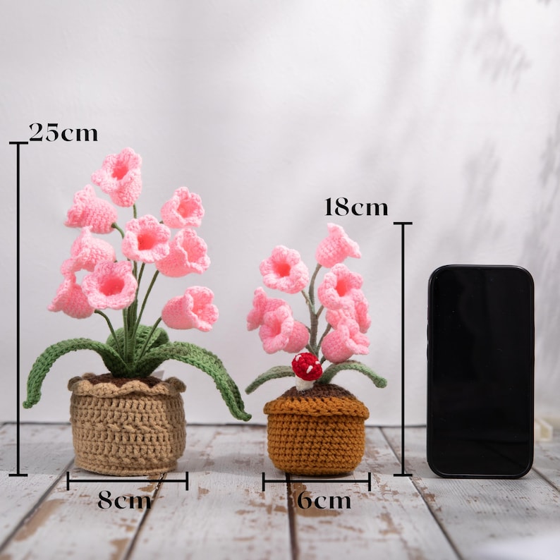 May include: Two crochet flower arrangements. The larger arrangement is 25cm tall and 8cm wide, featuring pink bell-shaped flowers in a tan pot. The smaller arrangement is 18cm tall and 6cm wide, with a red mushroom accent.